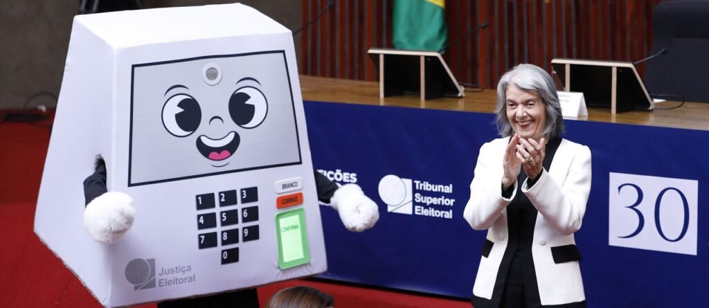 Mascote da urna eletronica ao lado de ministra do TSE, em evento oficial dos 30 anos da urna